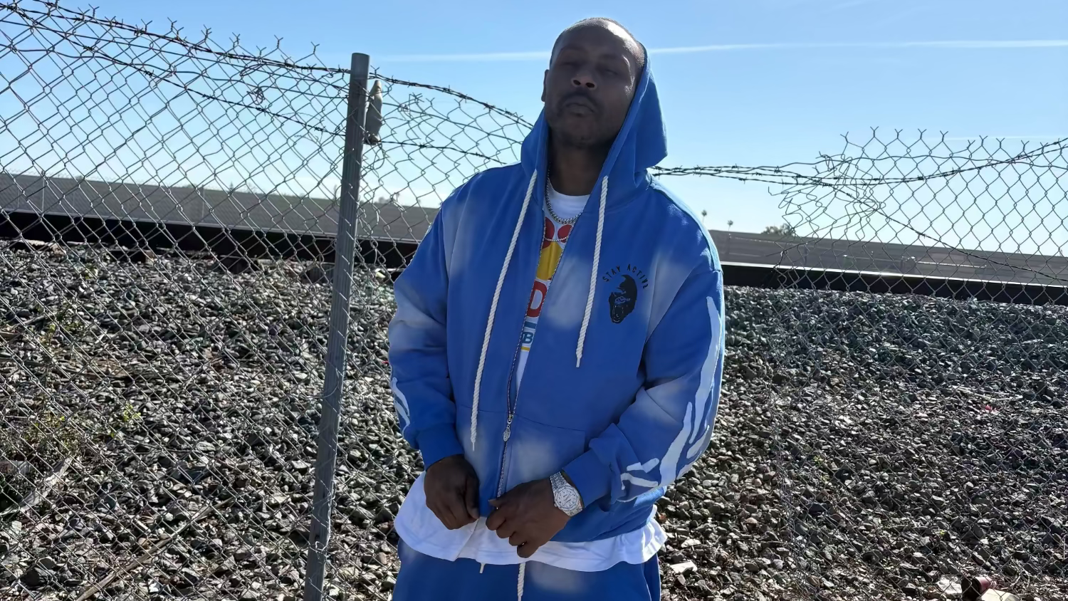 Person wearing a blue hoodie and pants standing in an outdoor area with a fence and clear sky.